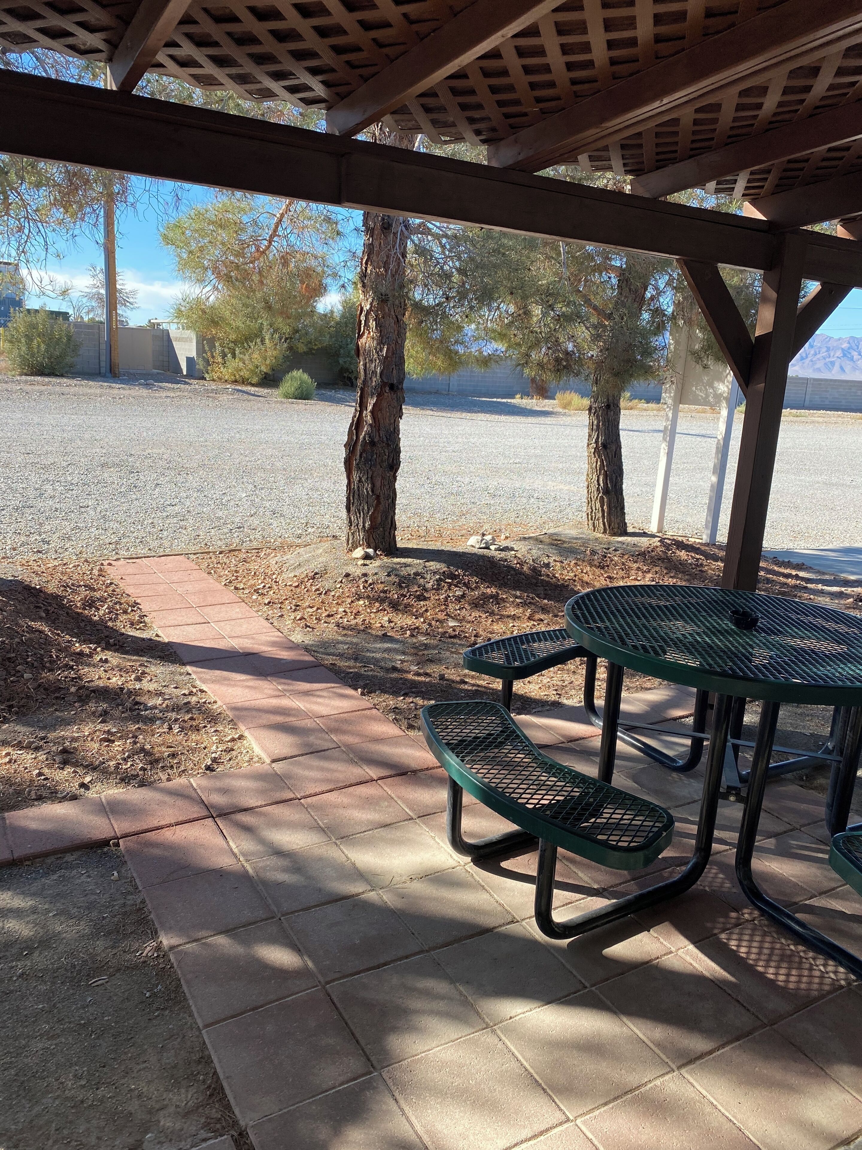 A covered picnic area with metal tables and benches, surrounded by gravel and trees.