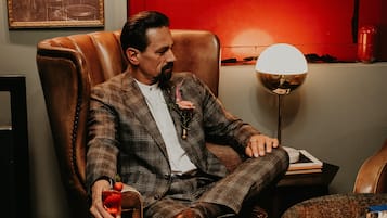 A man in a plaid suit sitting in a leather armchair with a drink in hand, a lamp, and a framed picture on the wall.