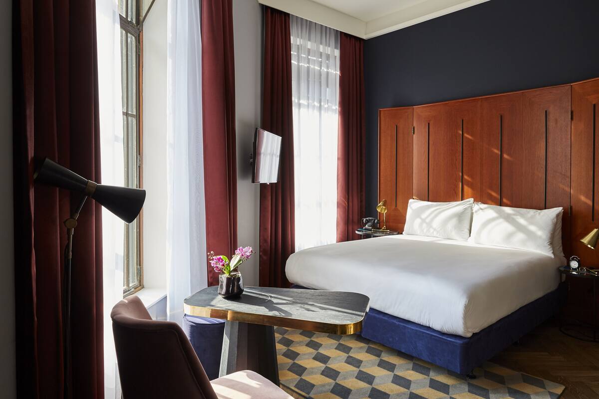 A hotel room with a large bed, a desk with a vase of flowers, a TV mounted on the wall, and a wooden headboard.