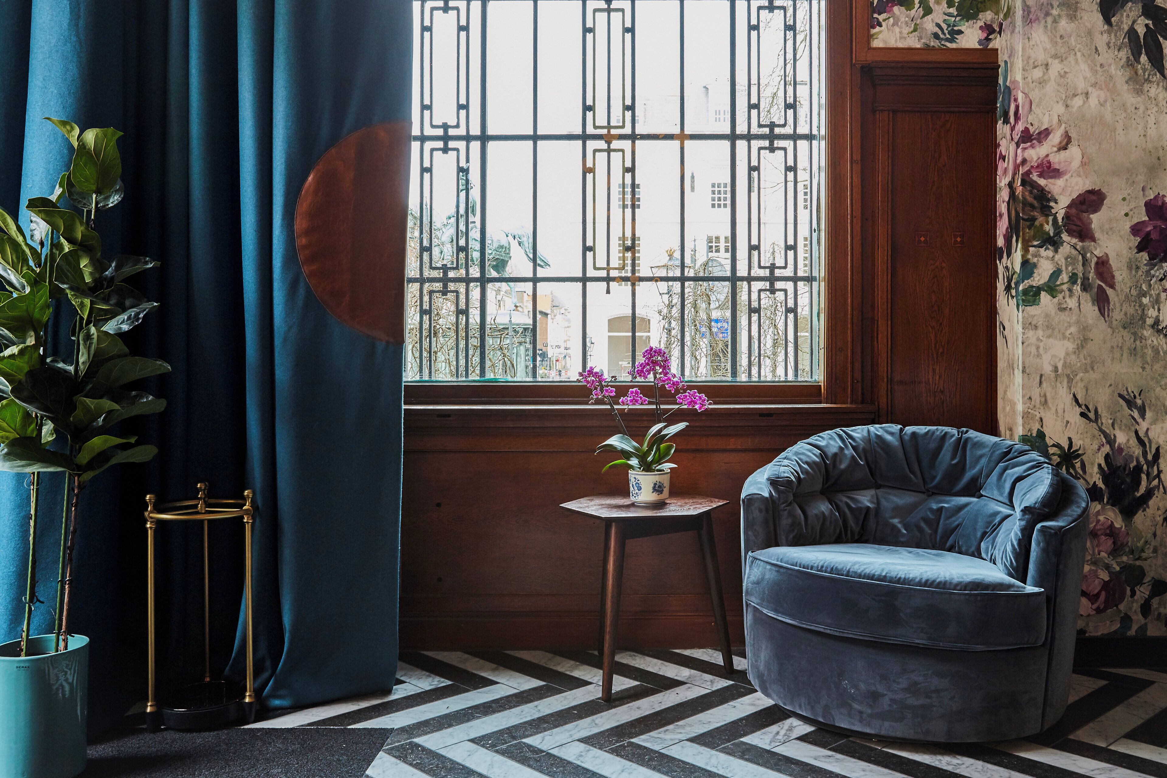 A corner room with a blue velvet armchair, a small table with a potted plant, and a window with a view of buildings.