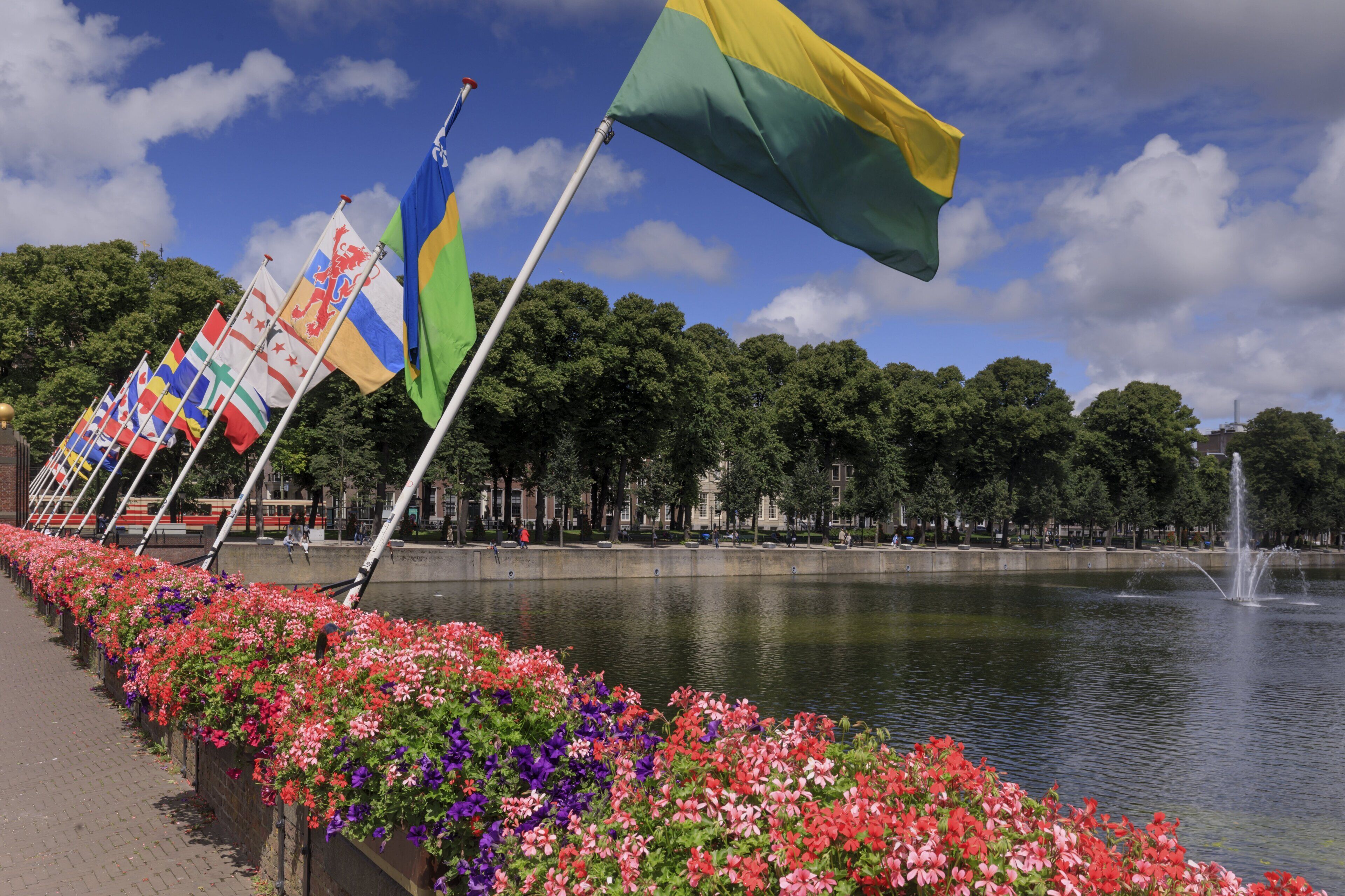 A waterfront promenade with flags, a fountain, and flower-lined path.