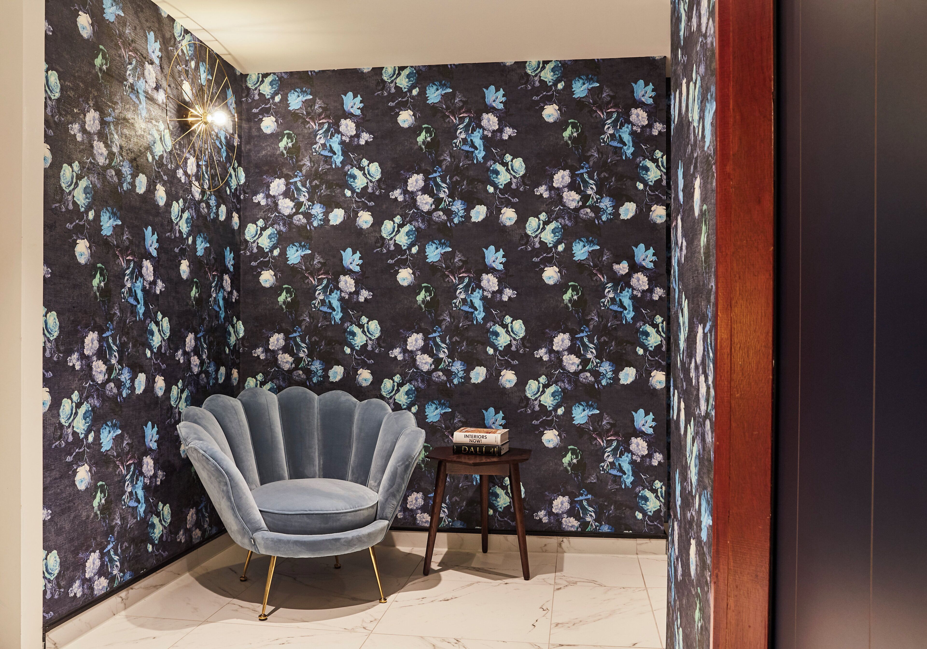 A corner room with a floral wallpaper, a blue upholstered chair, and a small wooden side table with books.