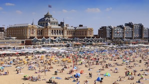 Ein weiter Strand mit vielen Sonnenschirmen und Menschen. Im Hintergrund ein historisches Gebäude mit einer Kuppel und Flaggen.