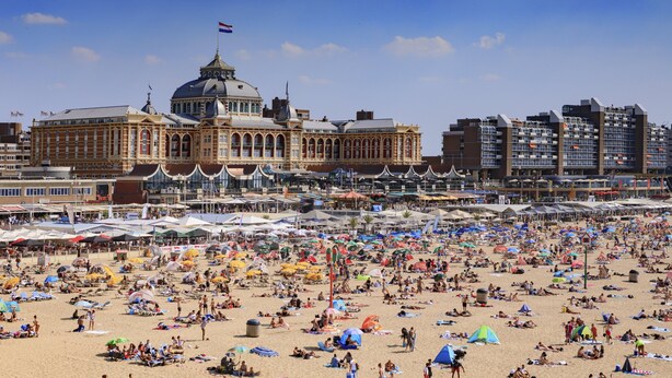 Une grande plage, avec de nombreux parasols et beaucoup de monde. Au fond, on aperçoit un bâtiment historique doté d’un dôme et de drapeaux.