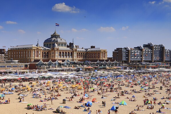Ein weiter Strand mit vielen Sonnenschirmen und Menschen. Im Hintergrund ein historisches Gebäude mit einer Kuppel und Flaggen.