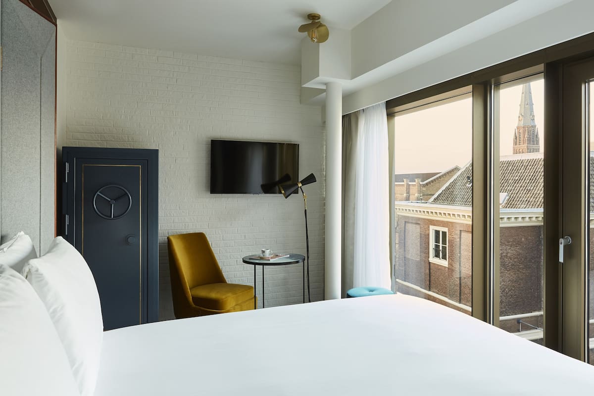 A hotel room with a large bed, a television mounted on the wall, a chair, a small table, and a view of a cityscape through large windows.