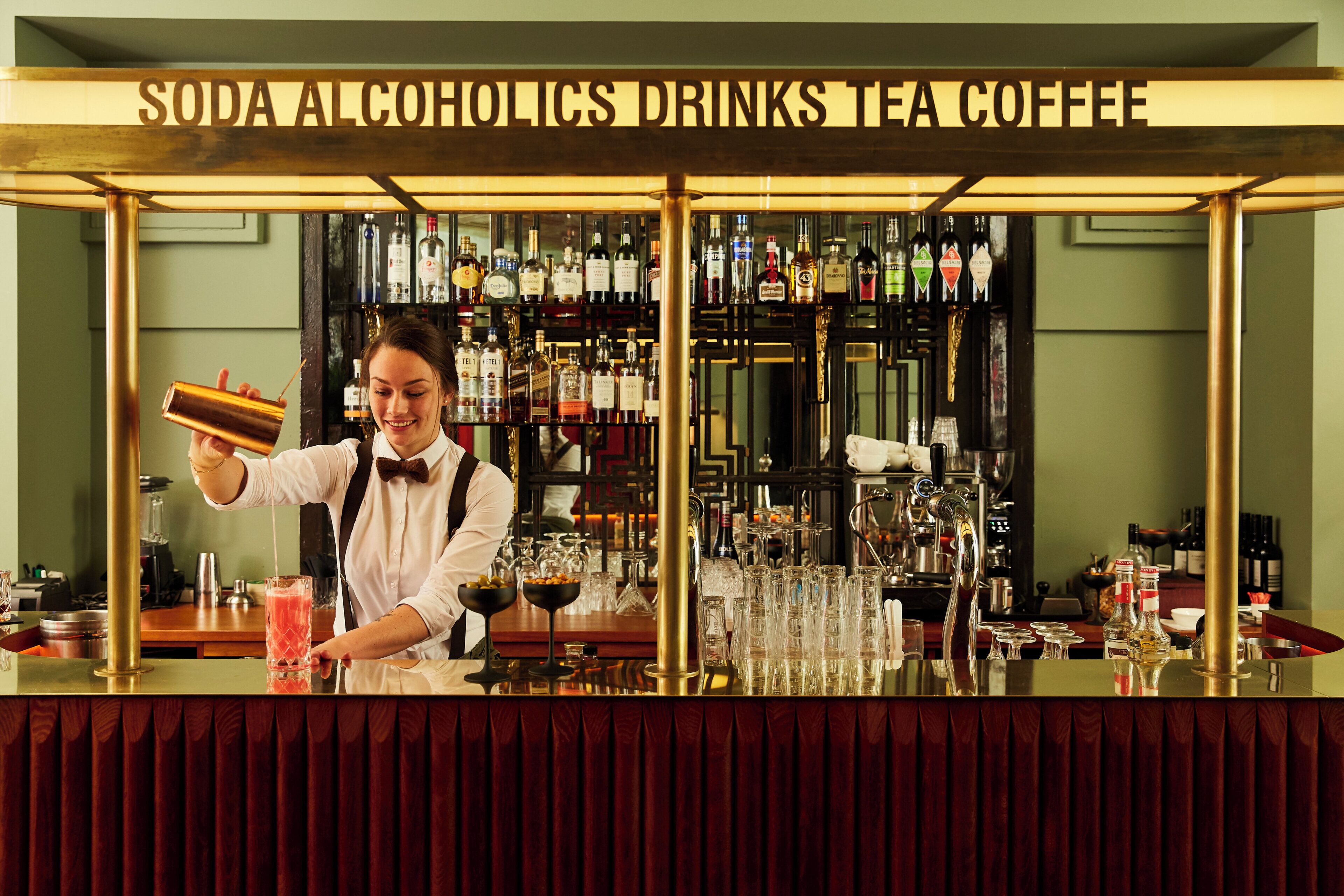 Zona de bar con un letrero que dice 'SODA ALCOHÓLICOS BEBIDAS TÉ CAFÉ' y un cantinero preparando una bebida.