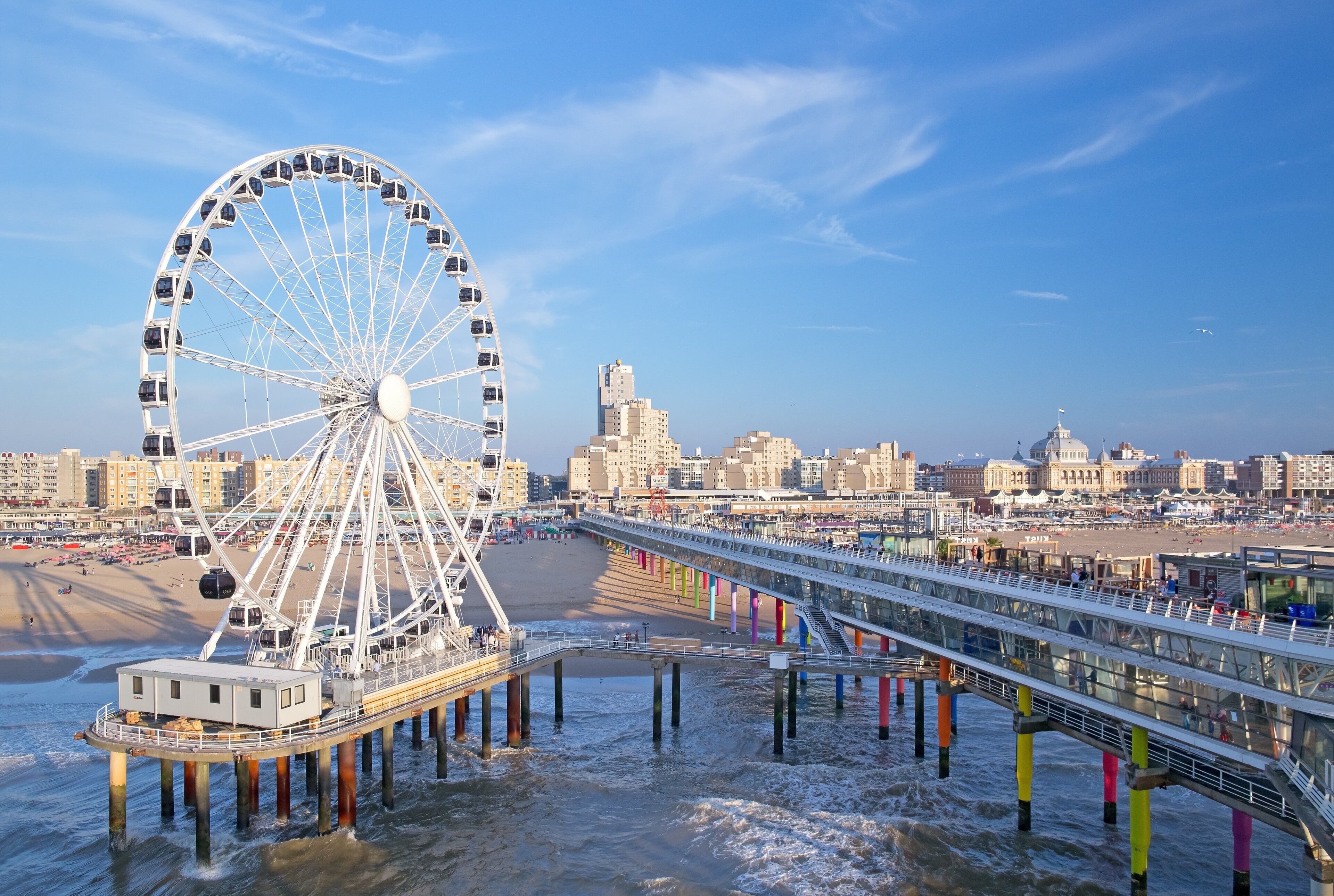 A Ferris wheel with a viewing platform, a pier extending into the water, and a cityscape in the background.