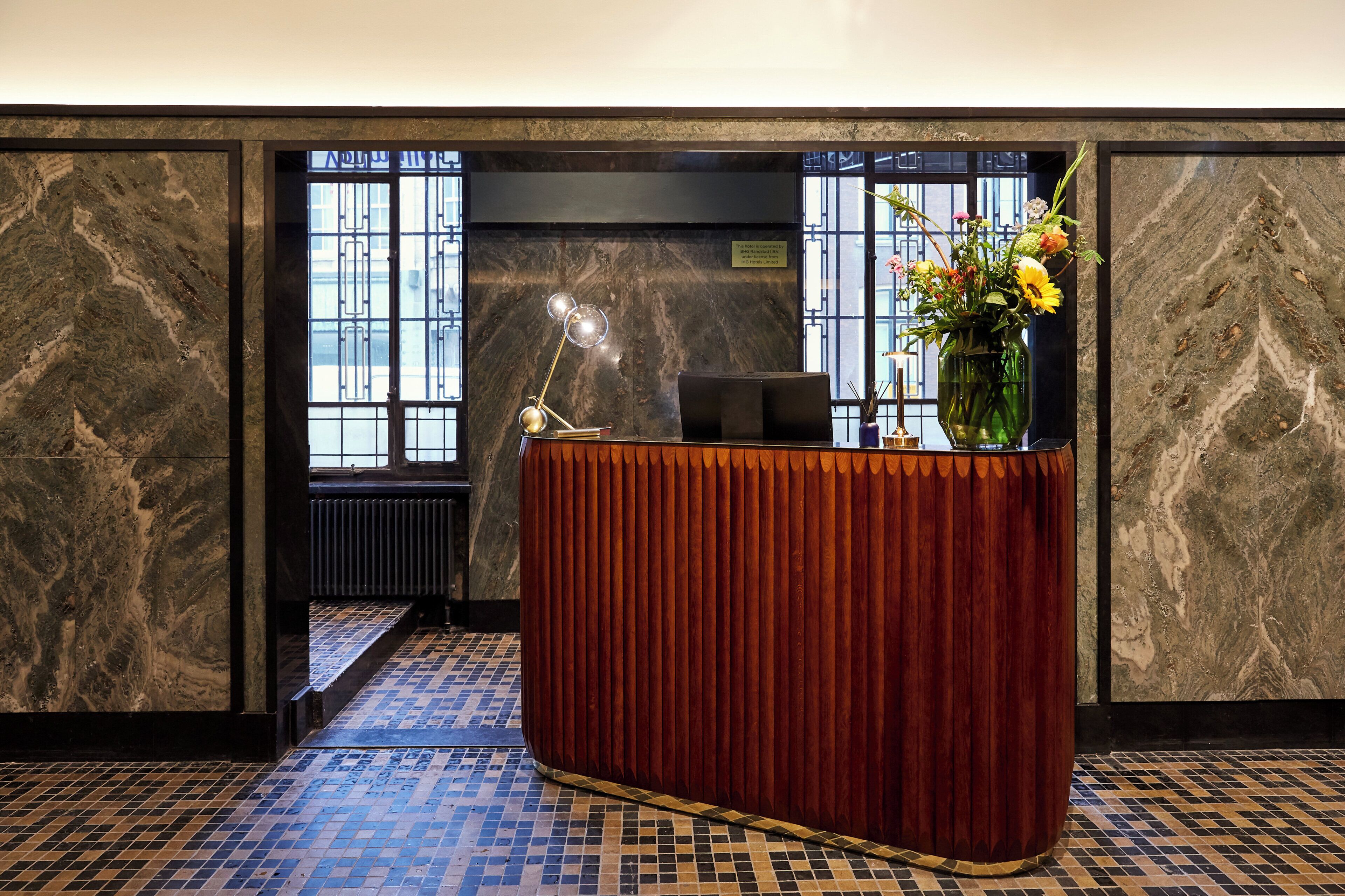 A reception desk with a vase of flowers, a desk lamp, and a view of the street through a window.