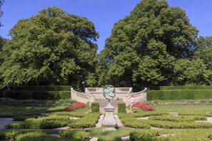 Ein formeller Garten mit einem zentralen Brunnen, gepflegten Rasenflächen und großen Bäumen.