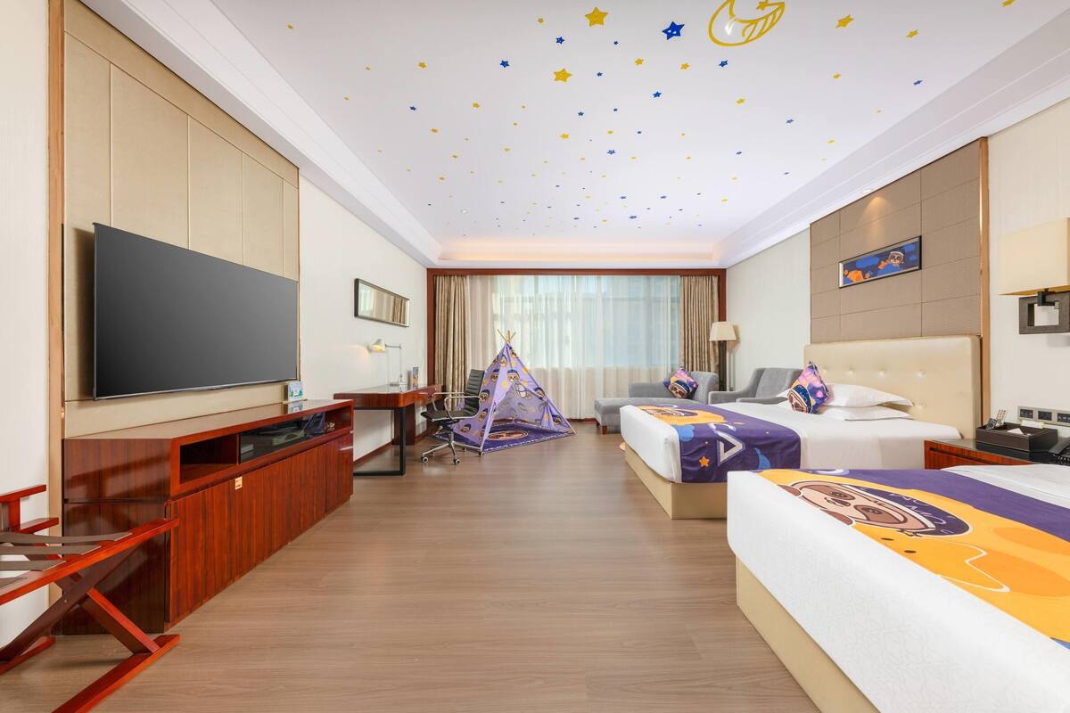 A hotel room with a large flat-screen TV, a desk, a chair, a bed with a colorful bedspread, a small tent, and a window with curtains.