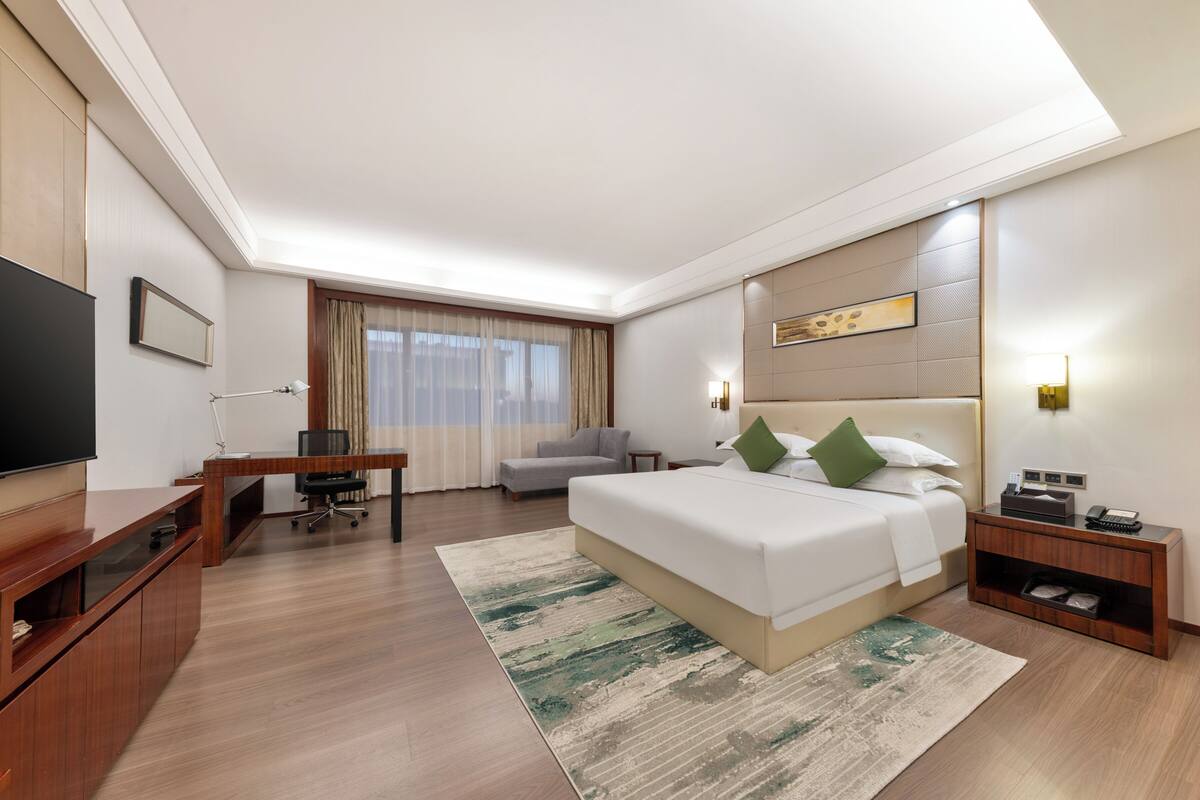 A modern hotel room with a large bed, a desk with a TV, a sofa, and a small table with a lamp.