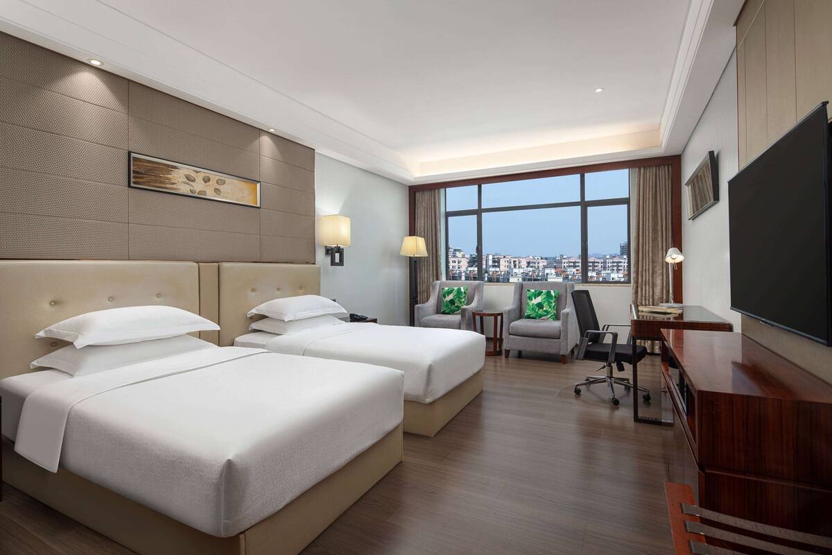 A hotel room with two beds, a desk, a chair, a TV, and a large window with a city view.