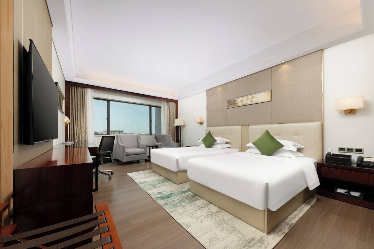 A hotel room with two beds, a desk, a TV, and a large window.