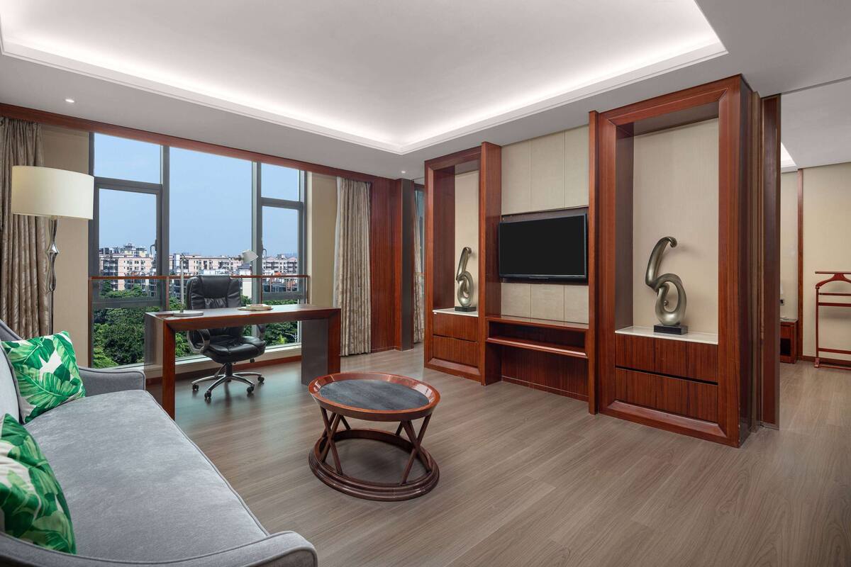 A modern hotel room with a large window, a sofa, a round coffee table, a desk, and a TV.