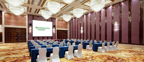A conference room with rows of tables and chairs, a large screen, and chandeliers.