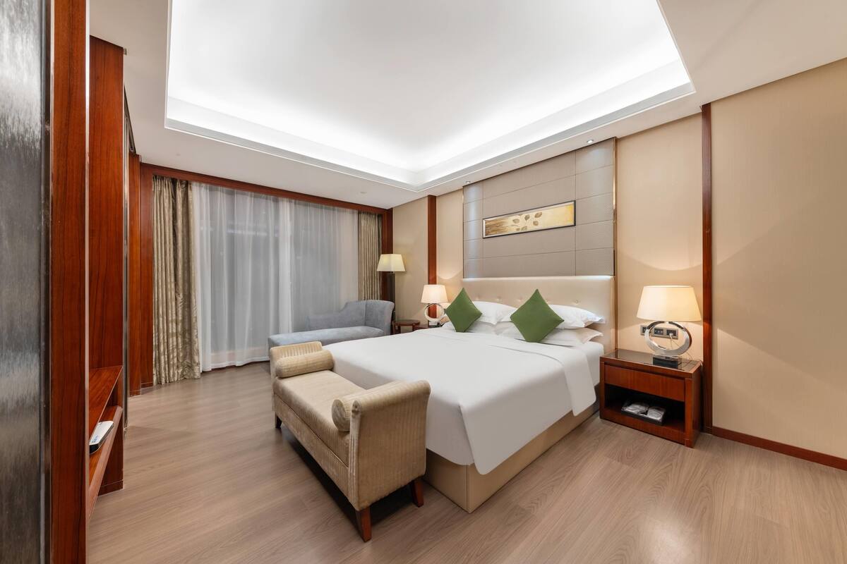 A hotel room with a large bed, a sofa, a wooden wardrobe, and a desk with a lamp.