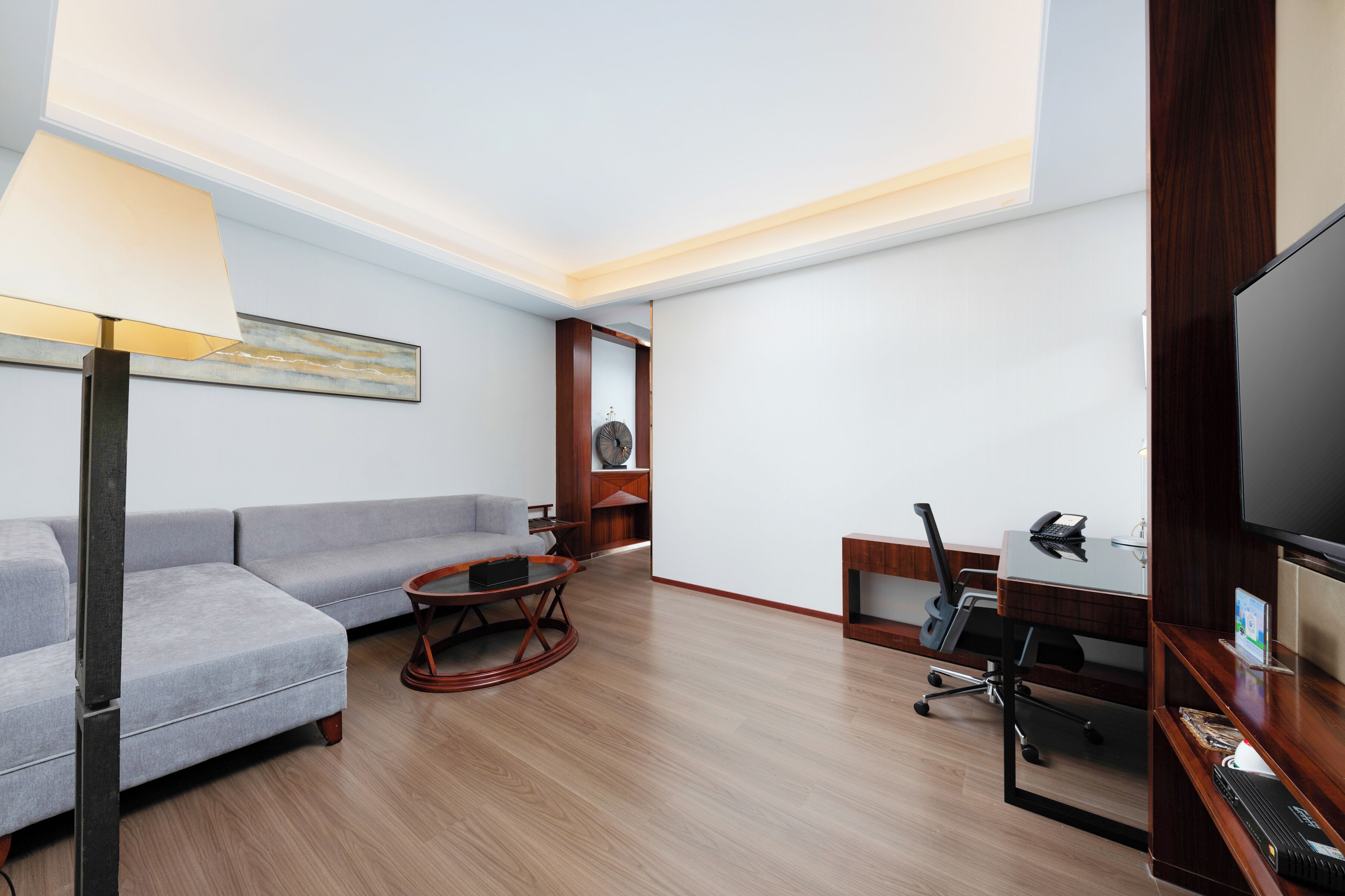 Habitación de hotel moderna con un sofá gris, una mesa de centro redonda de madera, un escritorio con una computadora y un televisor.