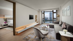 A modern living room with a sofa, armchair, and coffee table, a flat-screen TV mounted on the wall, and a large window offering a city view.