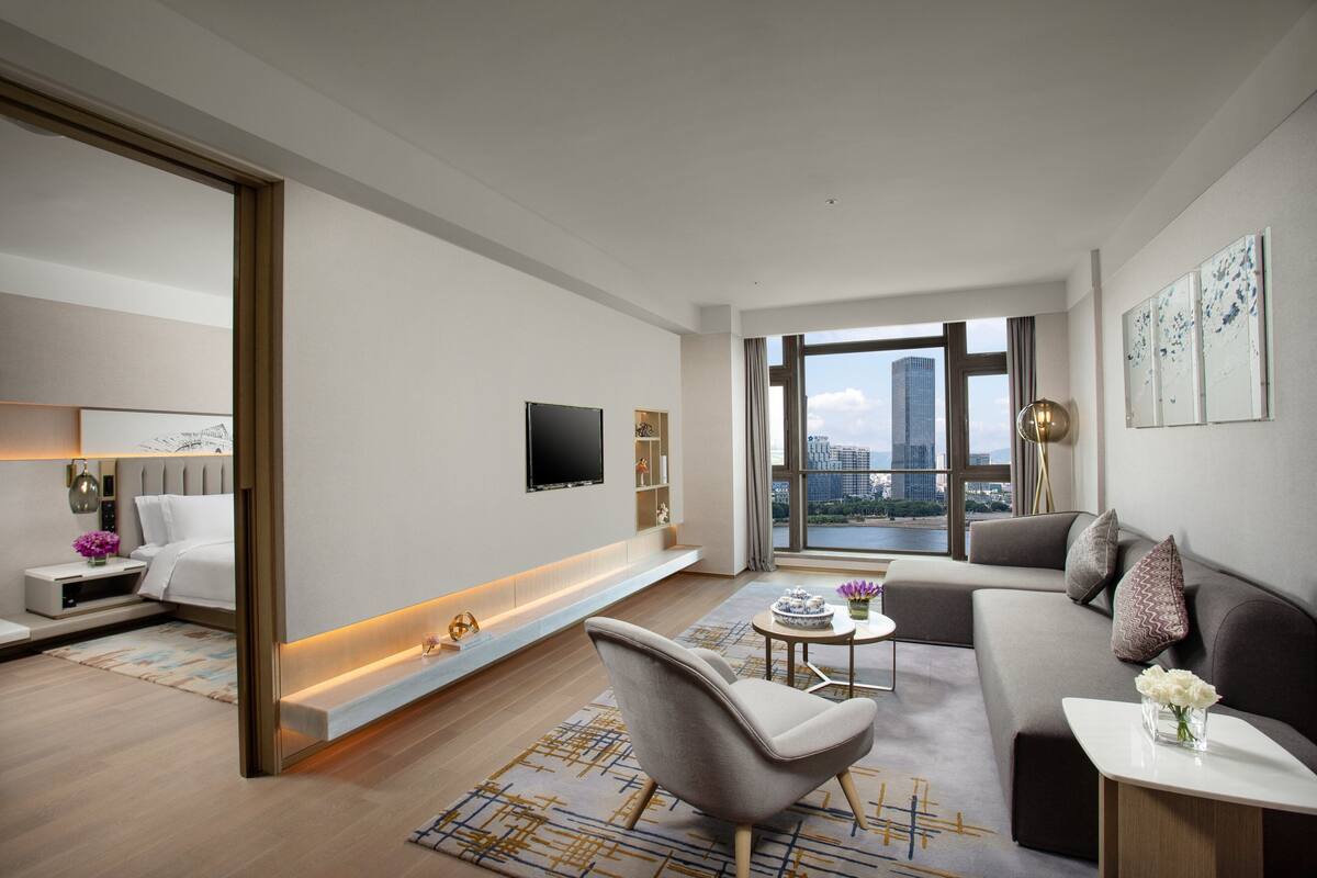 Ein modernes Wohnzimmer mit Sofa, Sessel und Couchtisch. Ein Flachbildfernseher ist an der Wand befestigt, und ein großes Fenster bietet Blick auf die Stadt.