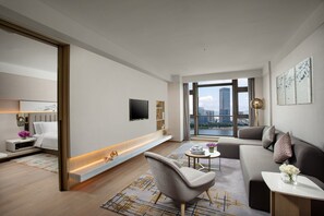 Una sala de estar moderna con sofá, sillón y mesa de centro, un televisor de pantalla plana montado en la pared, y un amplio ventanal con vista a la ciudad.