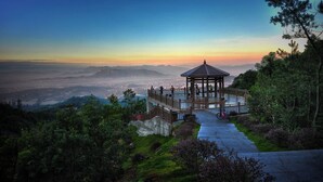 A gazebo with a wooden roof and railing, surrounded by lush greenery and a scenic view of distant mountains and a cityscape at sunset.