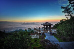 Un pabellón con techo y barandilla de madera, rodeado de exuberante follaje y una vista panorámica de montañas distantes y un paisaje urbano al atardecer.