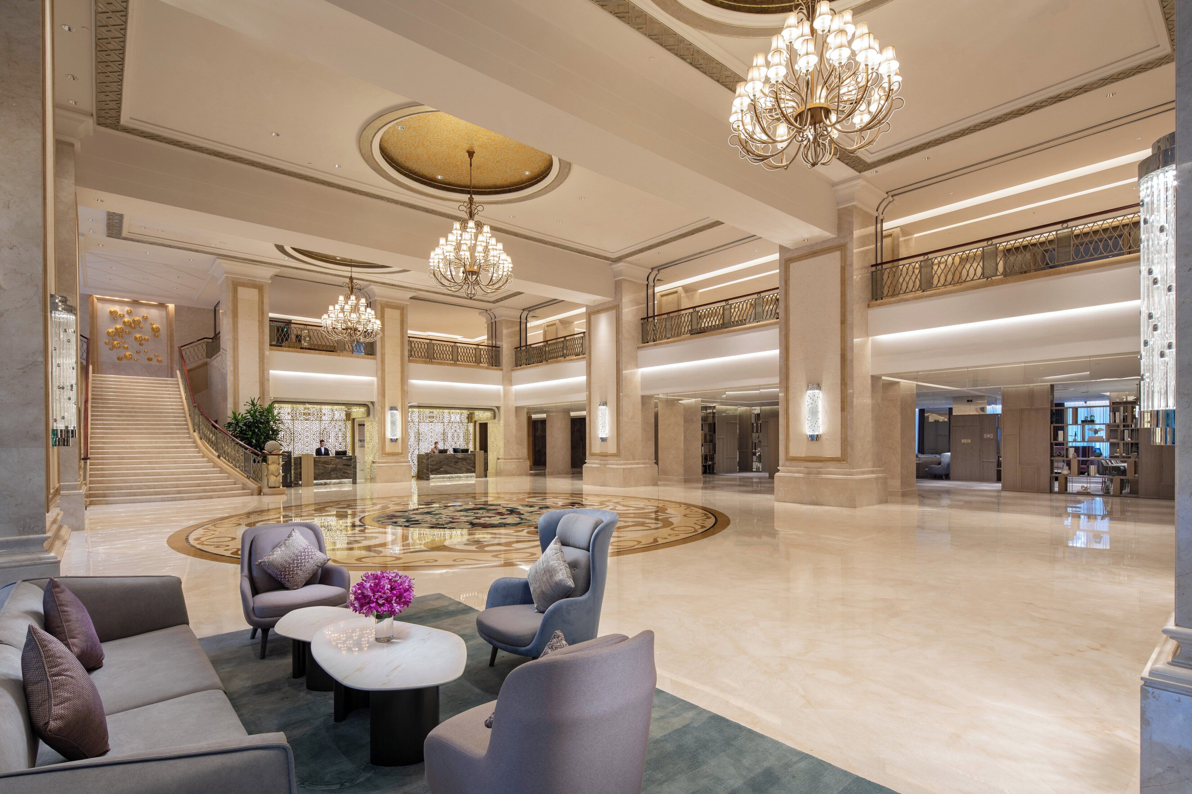 A spacious lobby with a grand staircase, multiple seating areas, and a central chandelier.