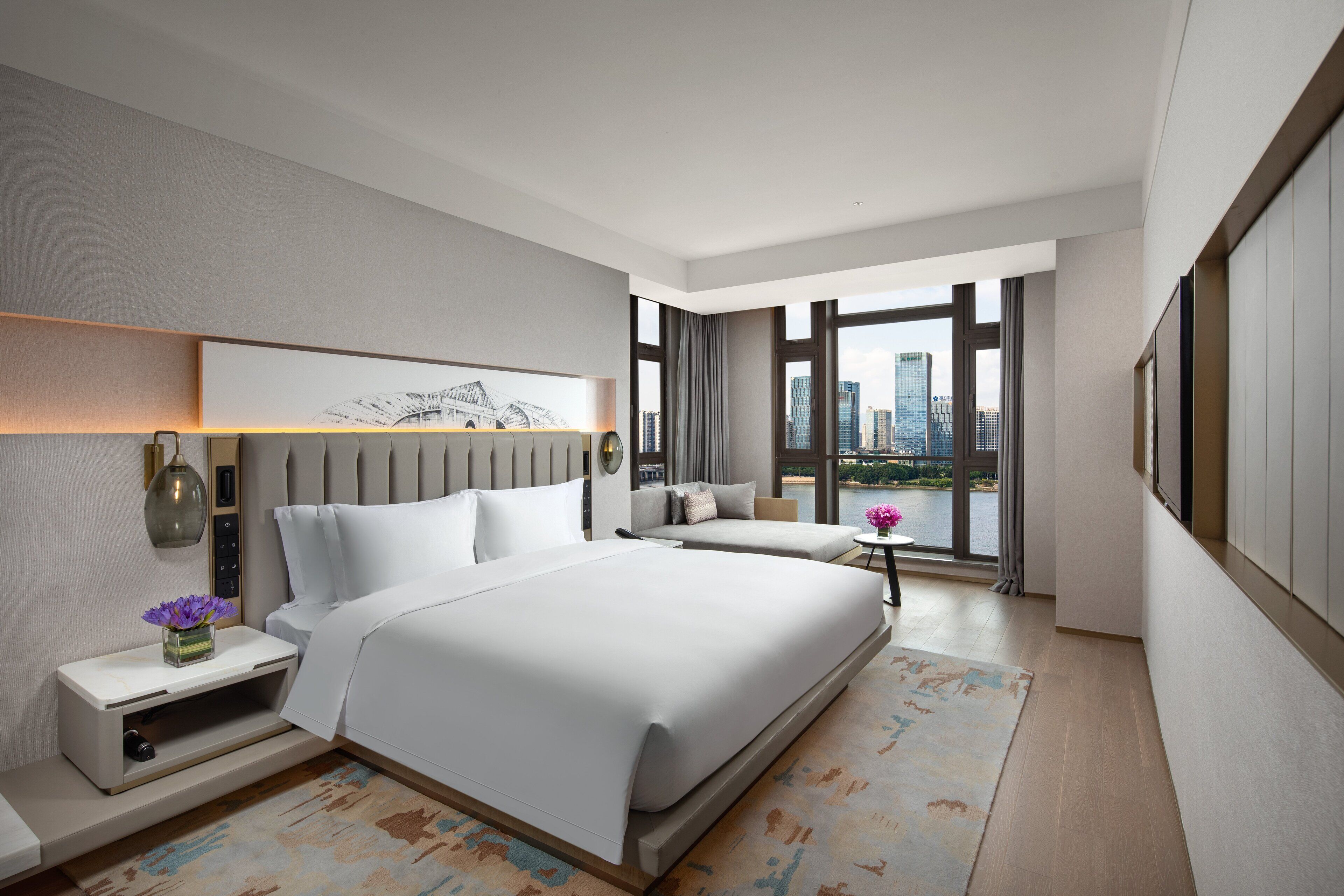 A modern hotel room with a large bed, a seating area, and a city view through large windows.
