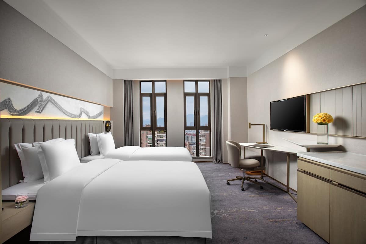 Ein Hotelzimmer mit zwei Betten, einem Schreibtisch, einem Sessel, einem Fernseher und einem großen Fenster mit Blick auf die Stadt.