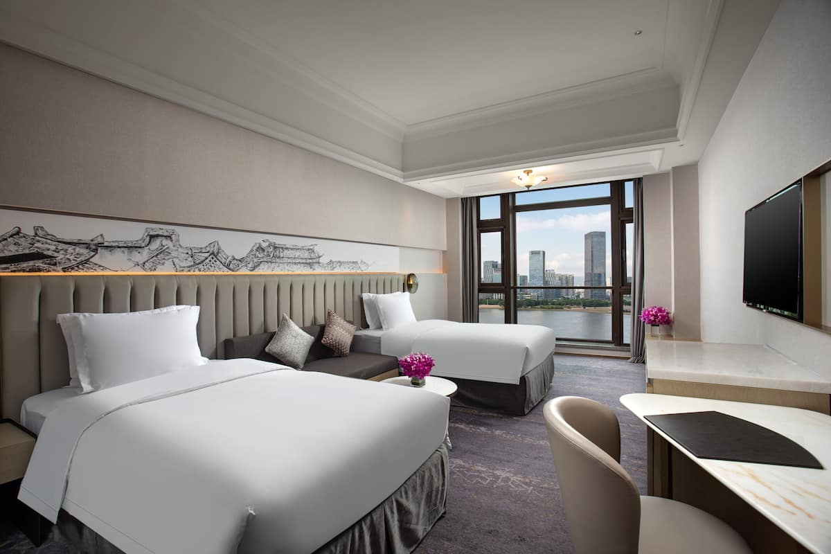 Ein Hotelzimmer mit zwei Betten, einem Schreibtisch und einem großen Fenster mit Blick auf die Stadt.
