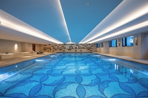 A large indoor swimming pool with a patterned floor, a reception area with seating, and a modern ceiling design.