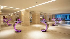 Un hall d’entrée spacieux, doté de tables modernes aux pieds incurvés et agrémenté d’éclairages violets, ainsi que d’un sol en marbre, et offrant une vue sur un bar situé au fond de la pièce.