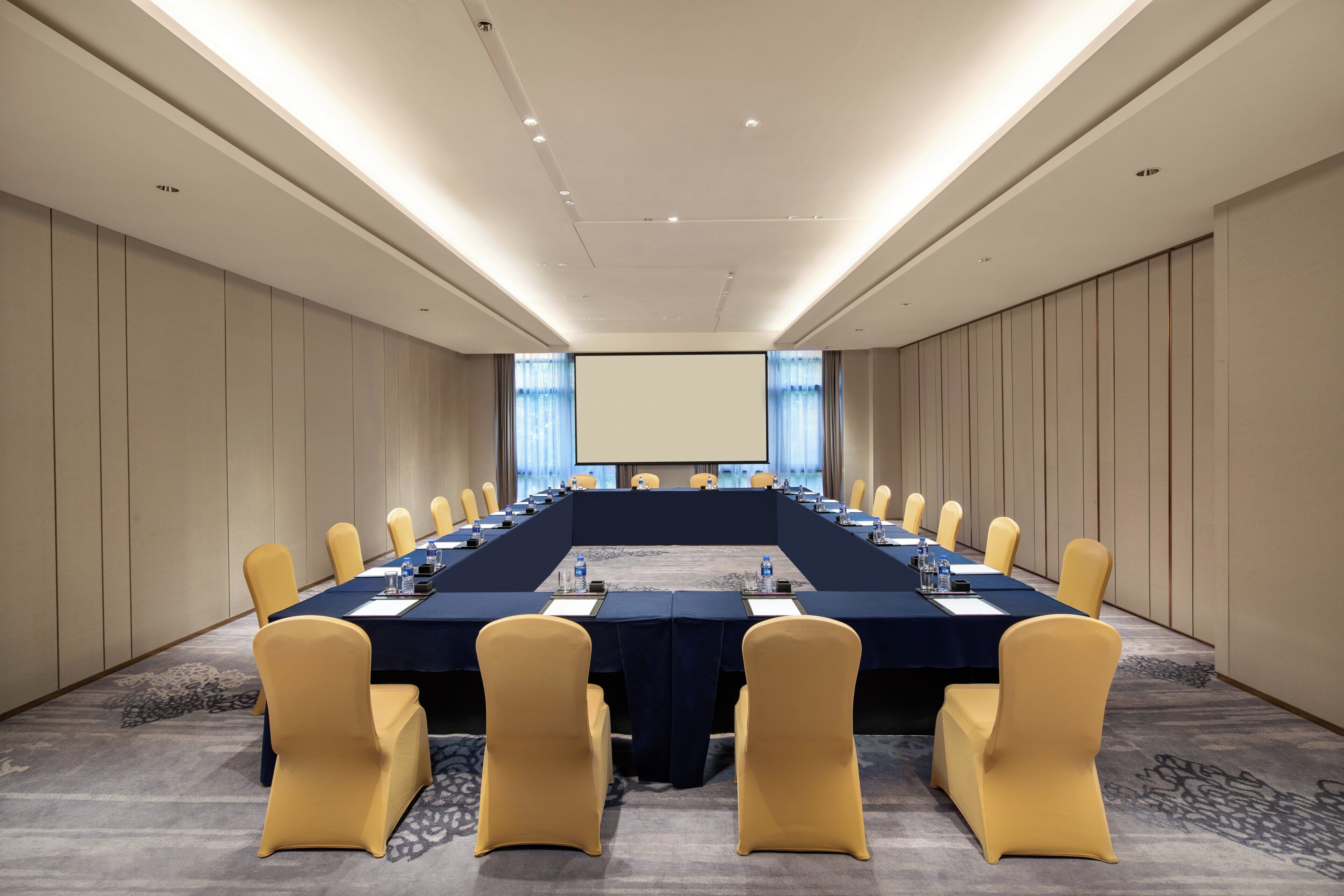 A conference room with a long table, chairs, a projector screen, and a carpeted floor.