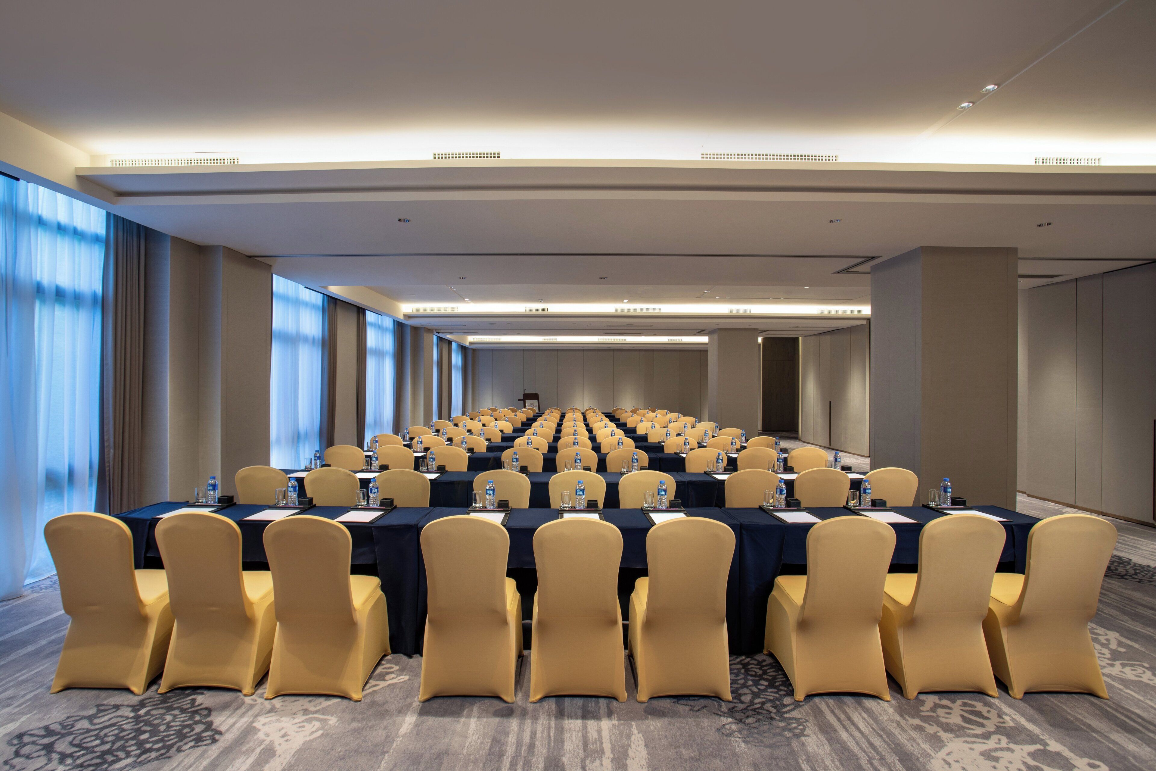 Una sala conferenze con file di tavoli e sedie, ampie finestre e pavimento rivestito di moquette.