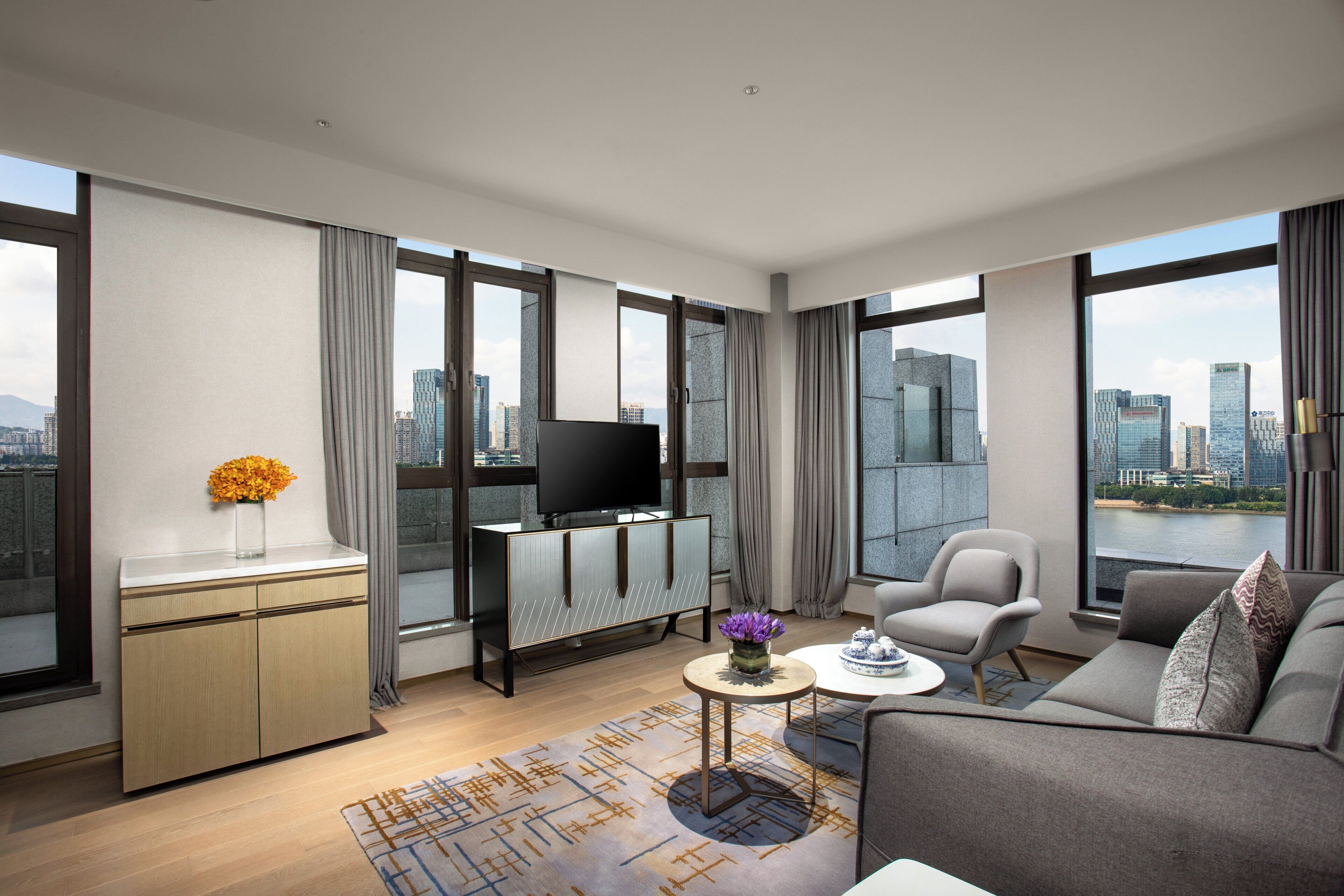 Un salon moderne avec un canapé, une table basse et un téléviseur. De grandes fenêtres offrent une vue sur la ville.