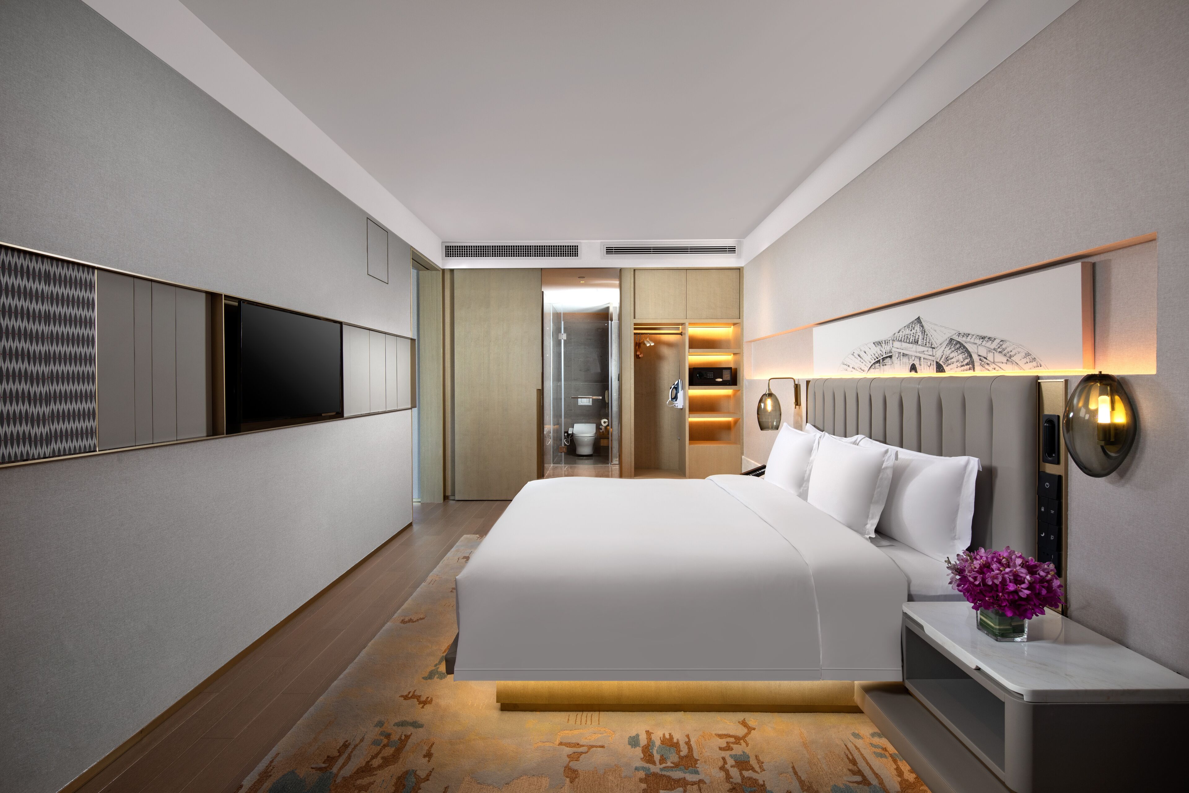 A modern hotel room with a large bed, a flat-screen TV mounted on the wall, a bedside table with a vase of flowers, and a walk-in closet.