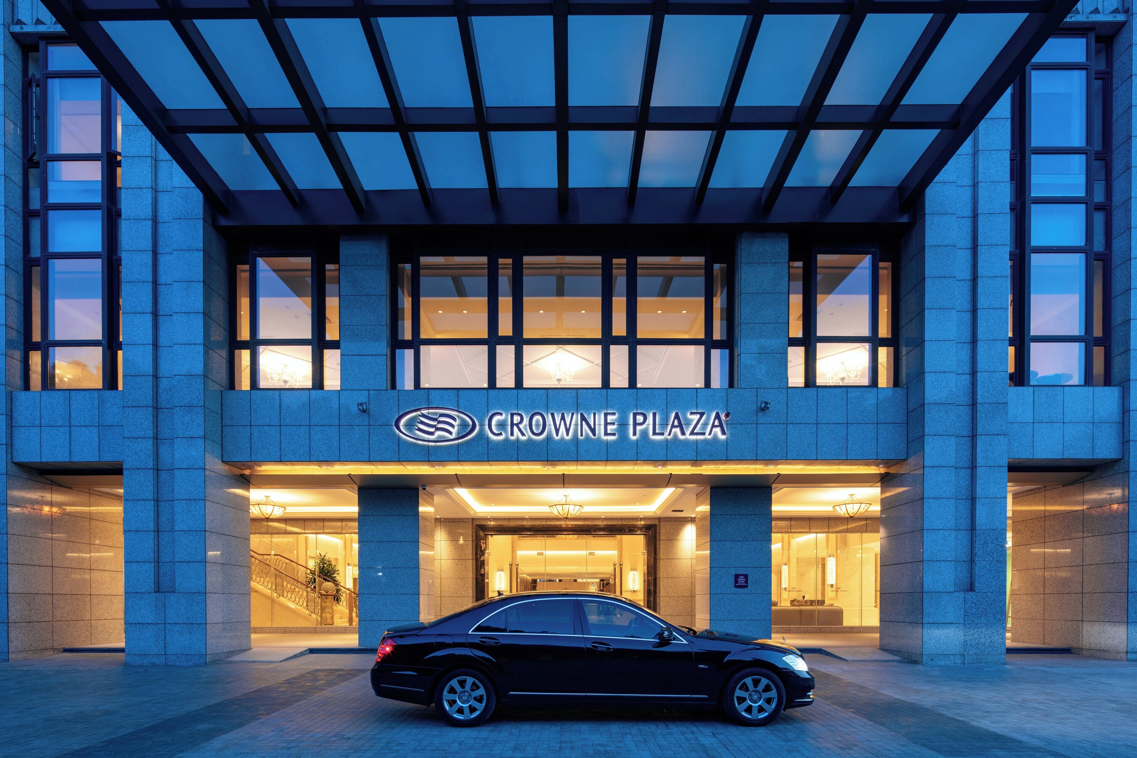 A hotel entrance with a car parked in front, a clear sky, and a sign reading 'CROWNE PLAZA'.