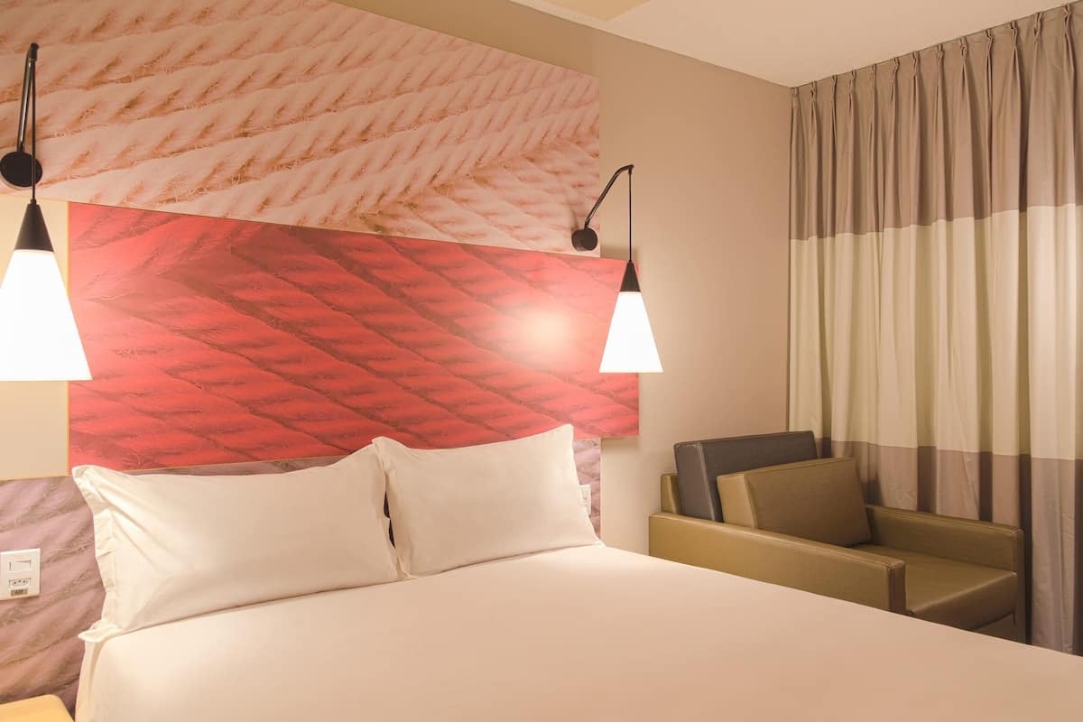 A hotel room with a bed, two bedside lamps, a headboard with a red patterned design, a sofa, and a curtain.