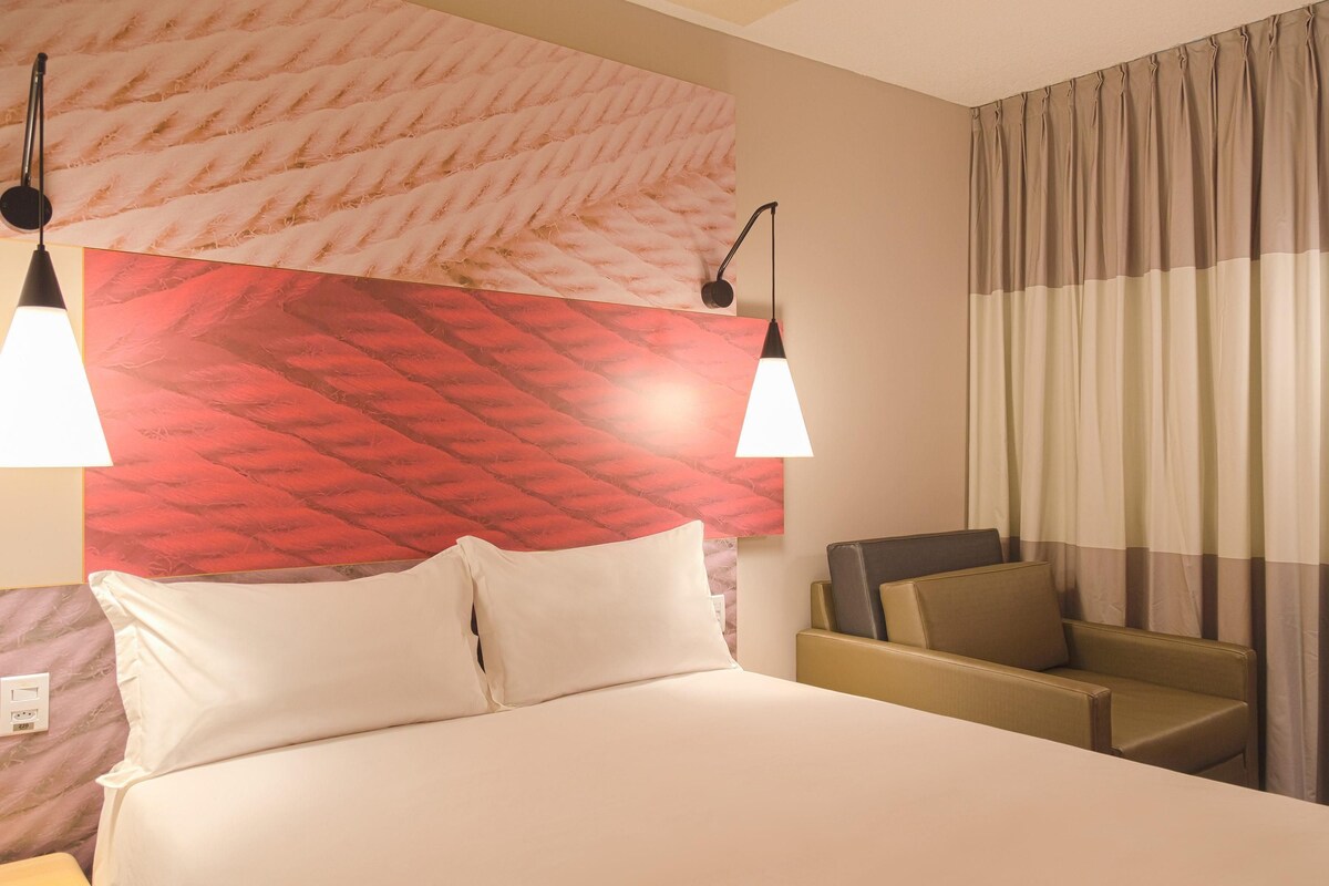 Habitación de hotel con cama, dos lámparas de noche, cabecera con diseño rojo estampado, sofá y cortina.