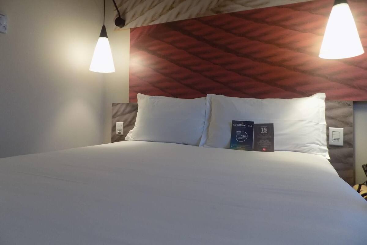 Ett hotellrum med en säng, två väggmonterade lampor, en röd sänggavel med mönster och en skylt som anger att incheckningen sker inom 15 minuter.