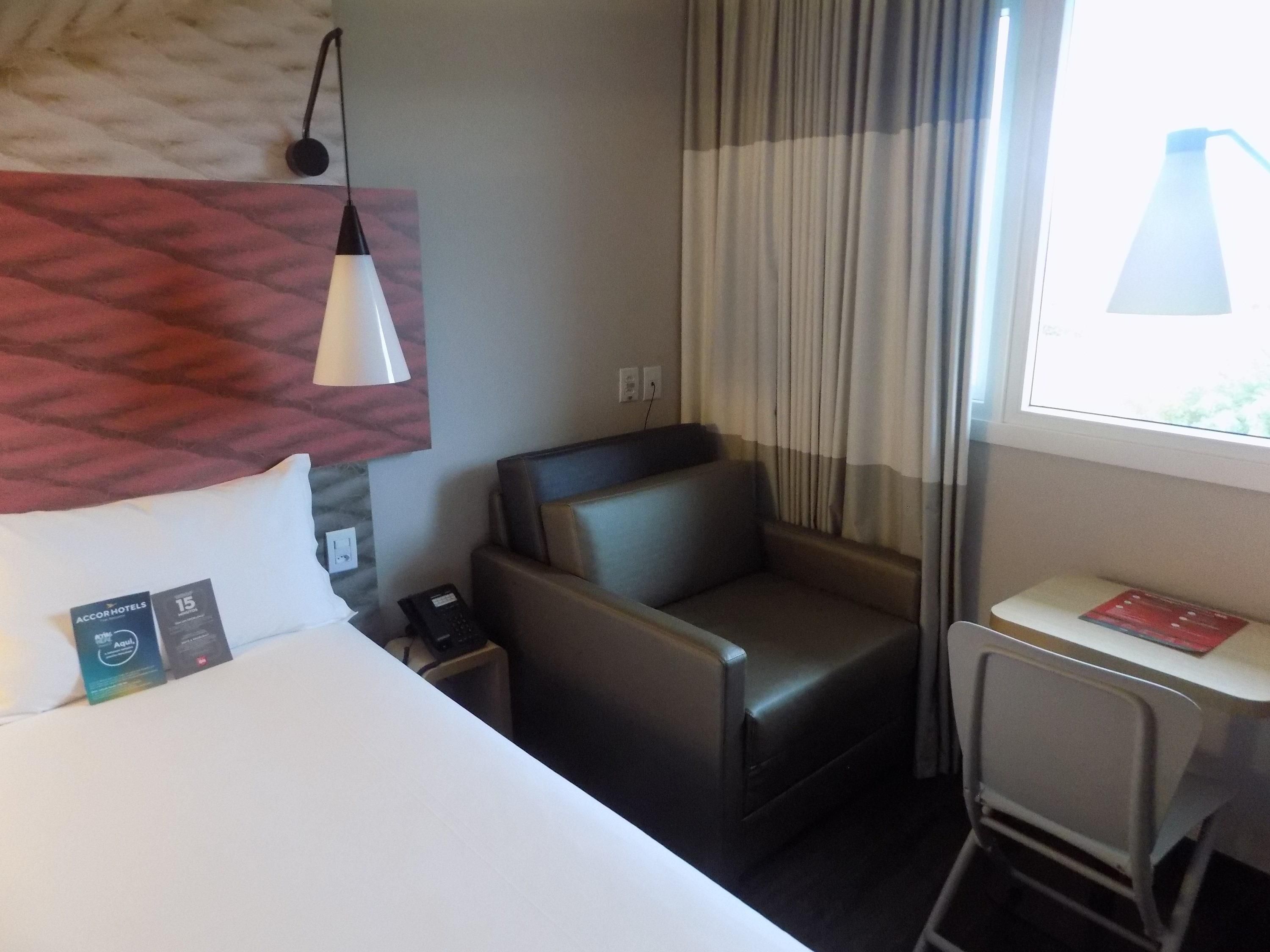 Una habitación de hotel con cama, escritorio, silla, teléfono y ventana con cortinas.