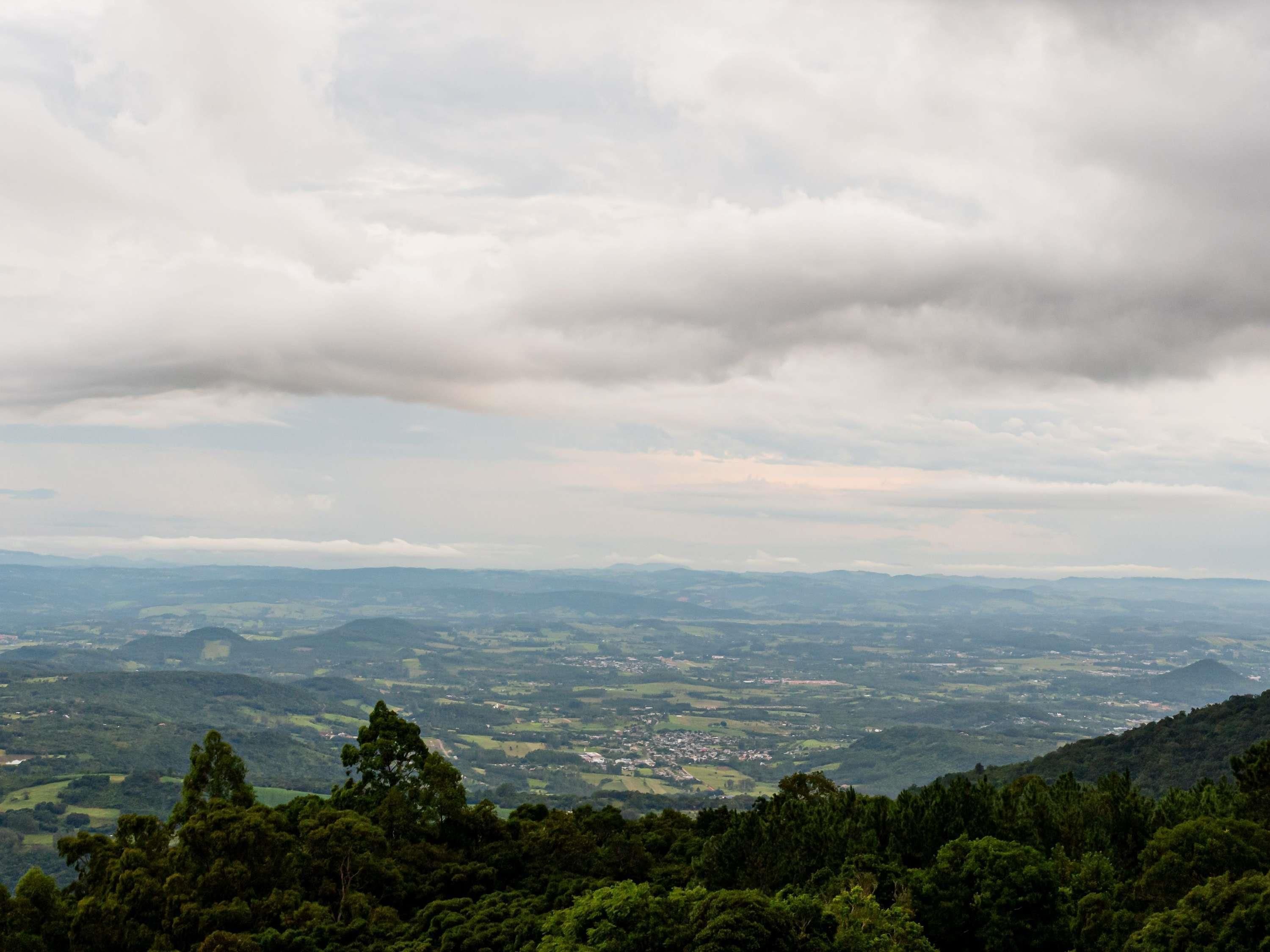 A panoramic view of a valley with a town in the distance, surrounded by dense forests and under a cloudy sky.