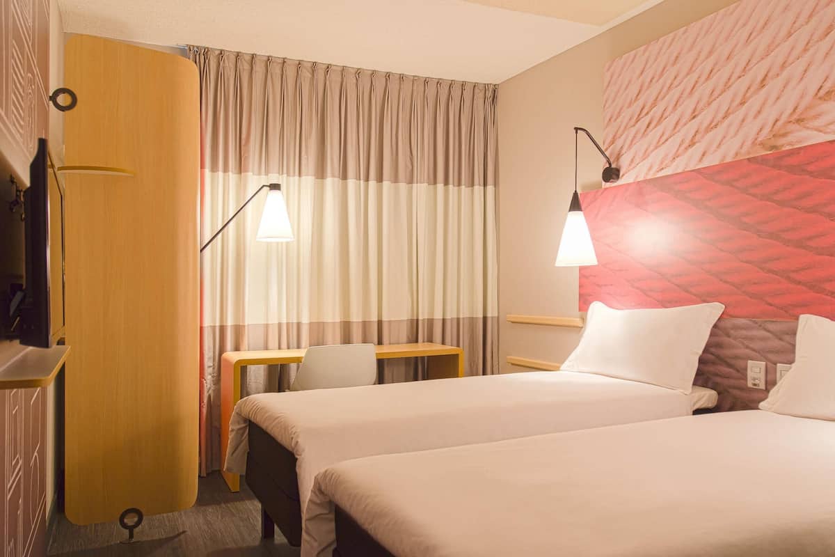 A hotel room with two beds, a desk, a chair, a lamp, and a curtain.