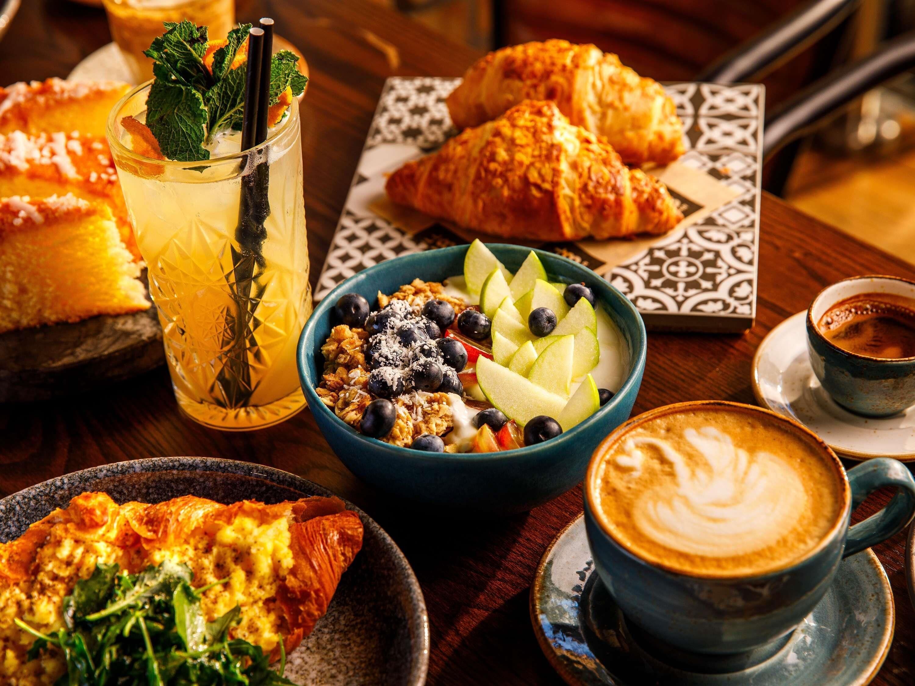 Une table sur laquelle on trouve une variété d’aliments, notamment des croissants, un saladier de fruits, un verre de boisson fraîche et une tasse de café.