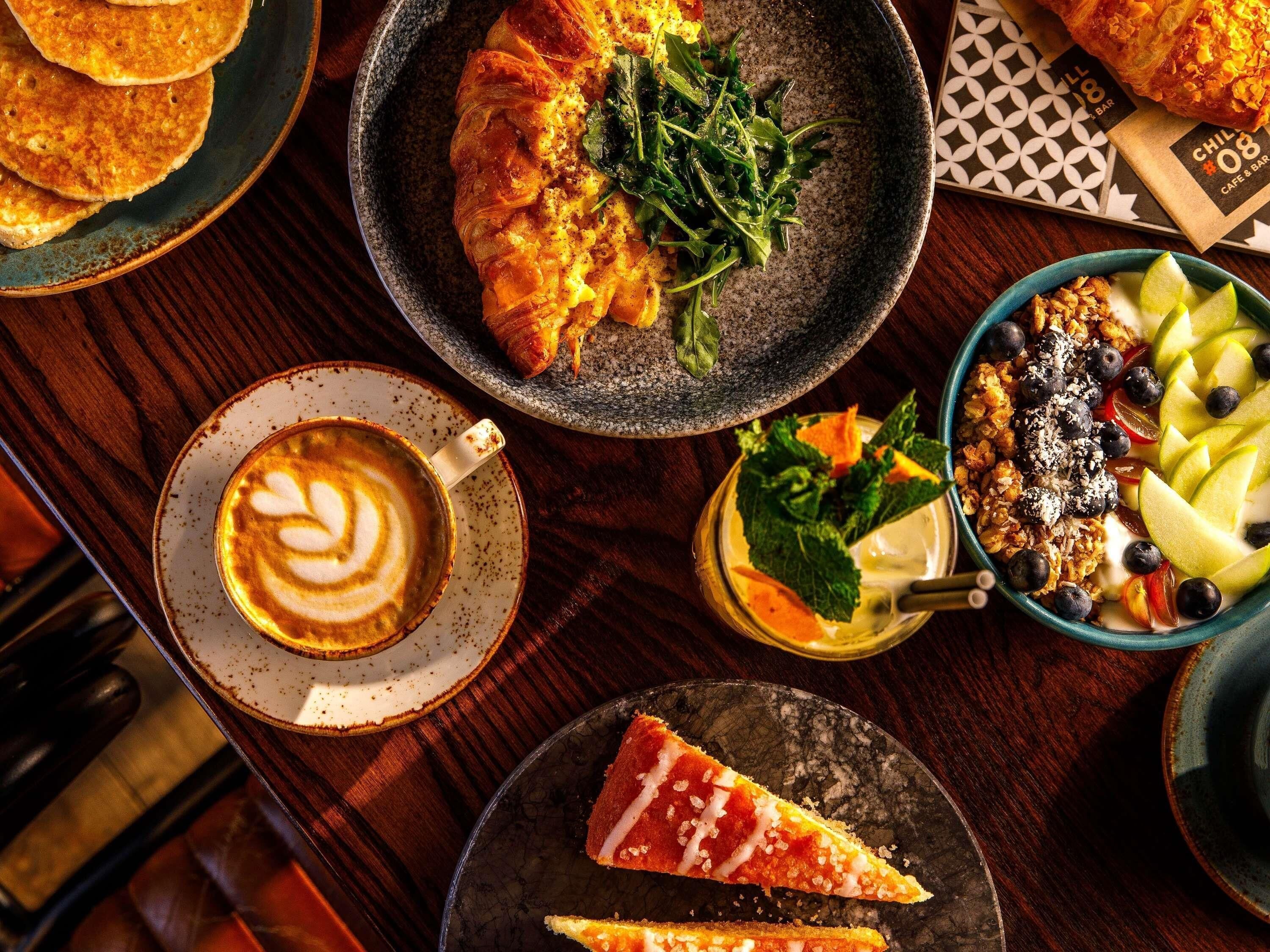 Variedad de alimentos para el desayuno, como croissants, un plato de ensalada y una porción de pizza, sobre una mesa de madera.