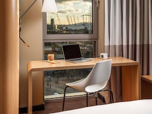 A modern hotel room with a wooden desk, a laptop, a coffee cup, and a view of a construction site through a window. - ibis London Canning Town (London)