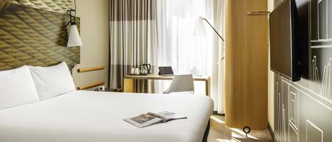 A hotel room with a large bed, a desk with a laptop, a television, and a window with curtains.