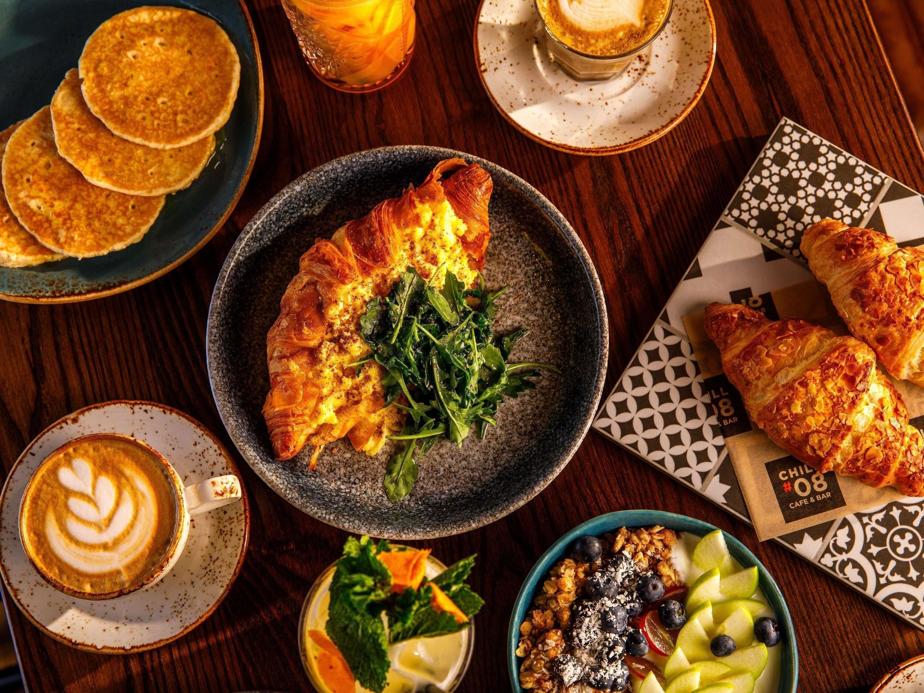 A breakfast setting with croissants, pancakes, a latte, and a bowl of fruit and granola.