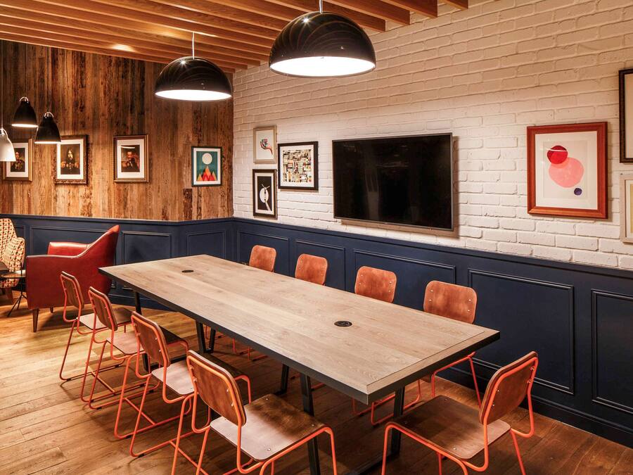 Una sala de conferencias con una larga mesa de madera y sillas anaranjadas, una pared de ladrillo blanco con obras de arte, y un televisor de pantalla plana grande.